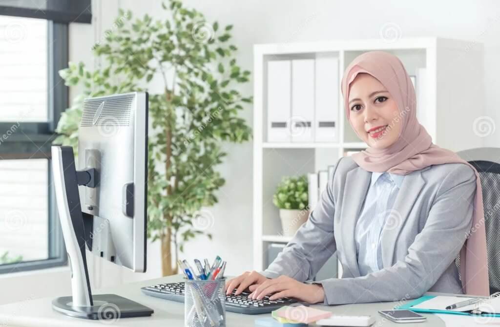 indonesian office lady sitting typing e mail face to camera smiling young female professional businesswoman working indoors 128547595